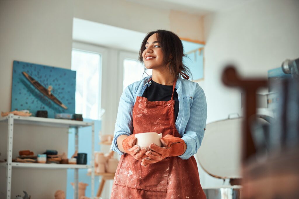 Cheerful female potter in apron holding handmade ceramic bowl