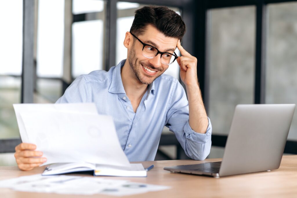 Happy businessman holding business documents, looking at laptop screen, smiling