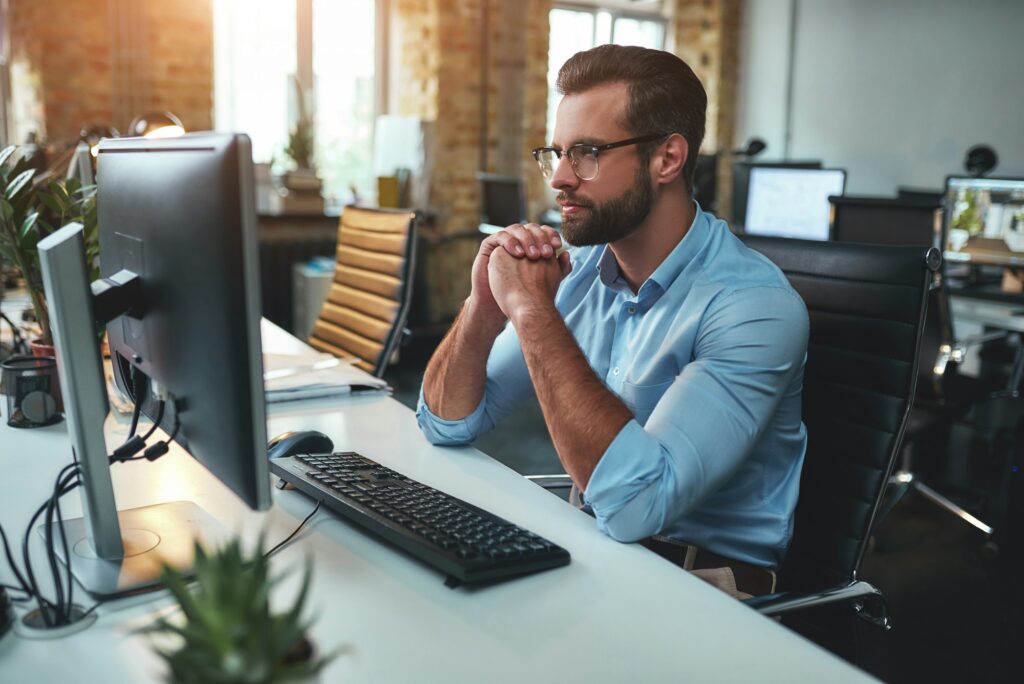 Thinking. Side view of young bearded man in eyeglasses and formal wear working on computer and